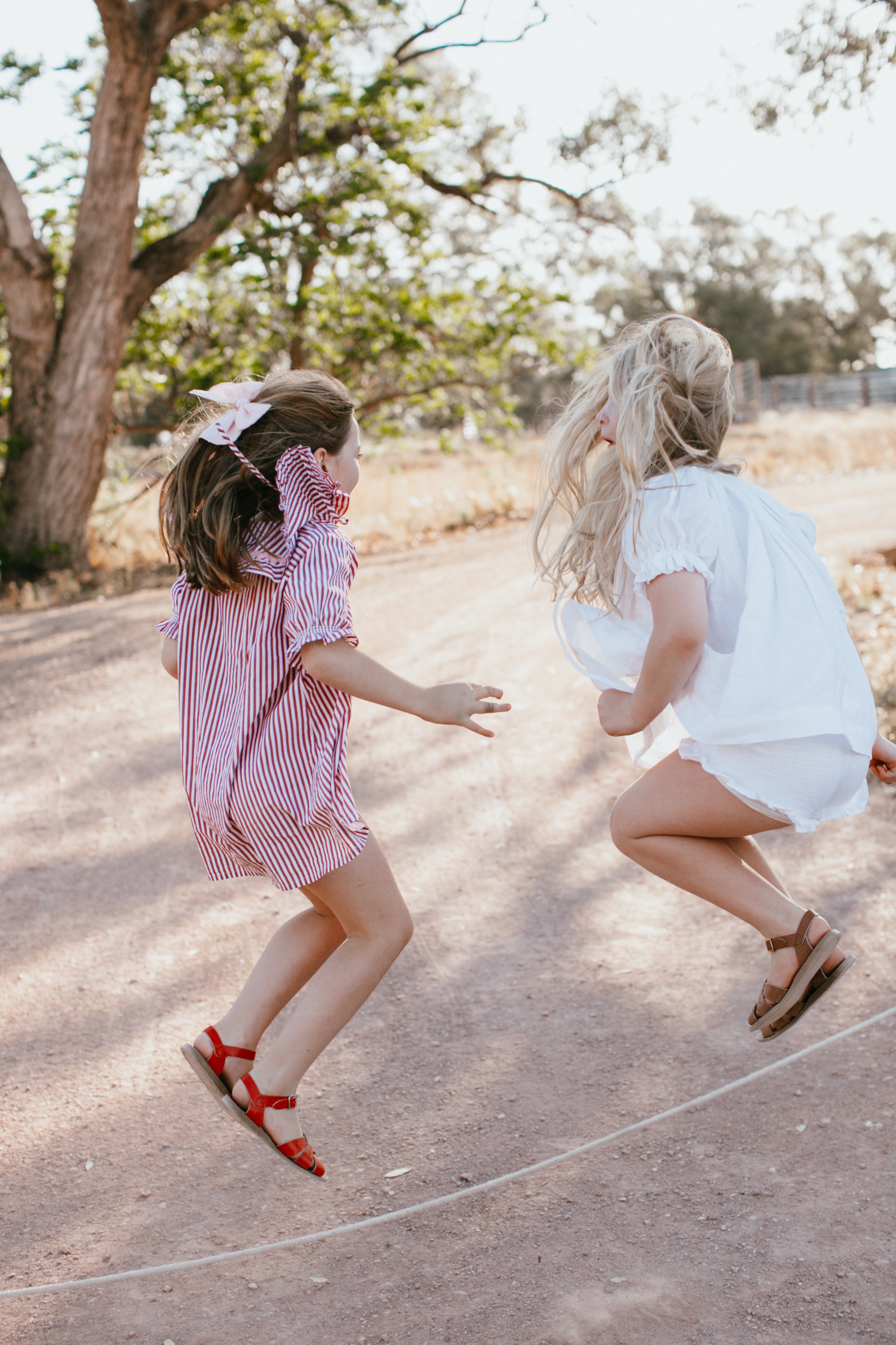 Two young girls jumping on a dirt path with trees in the background